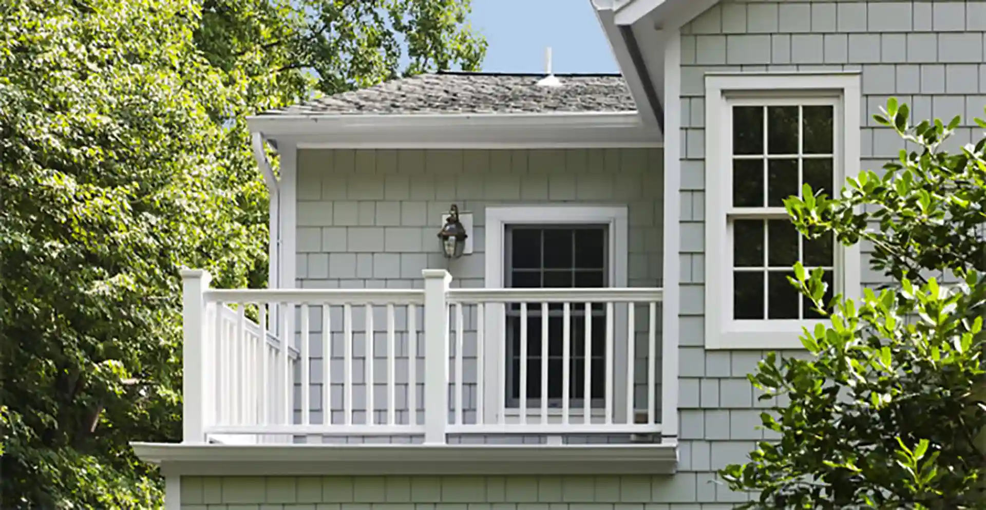 Small second-story balcony with white railing on a light gray shingle-sided home, surrounded by greenery.
