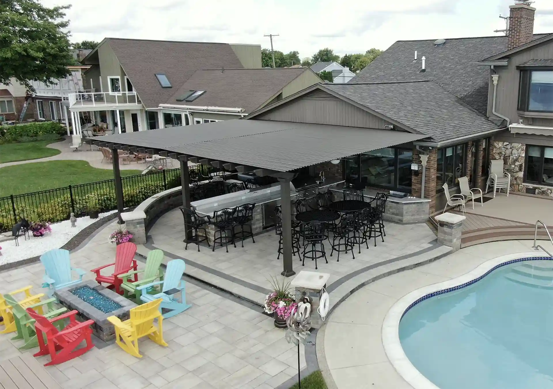 Outdoor patio with a large pergola-covered dining area, poolside seating, and a fire pit with colorful Adirondack chairs.
